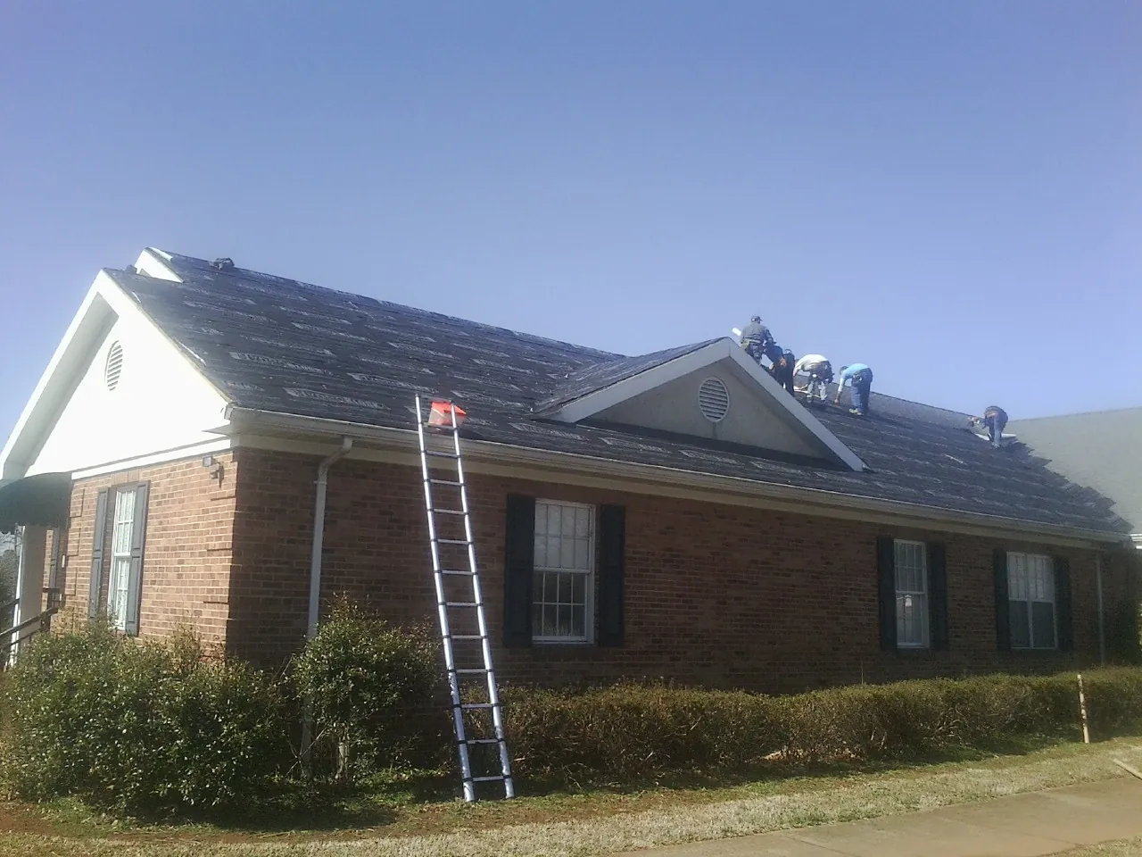 Crew tearing shingles off a brick ranch roof with a ladder set — Emergency Roof Repair work by our crew in Hayfield