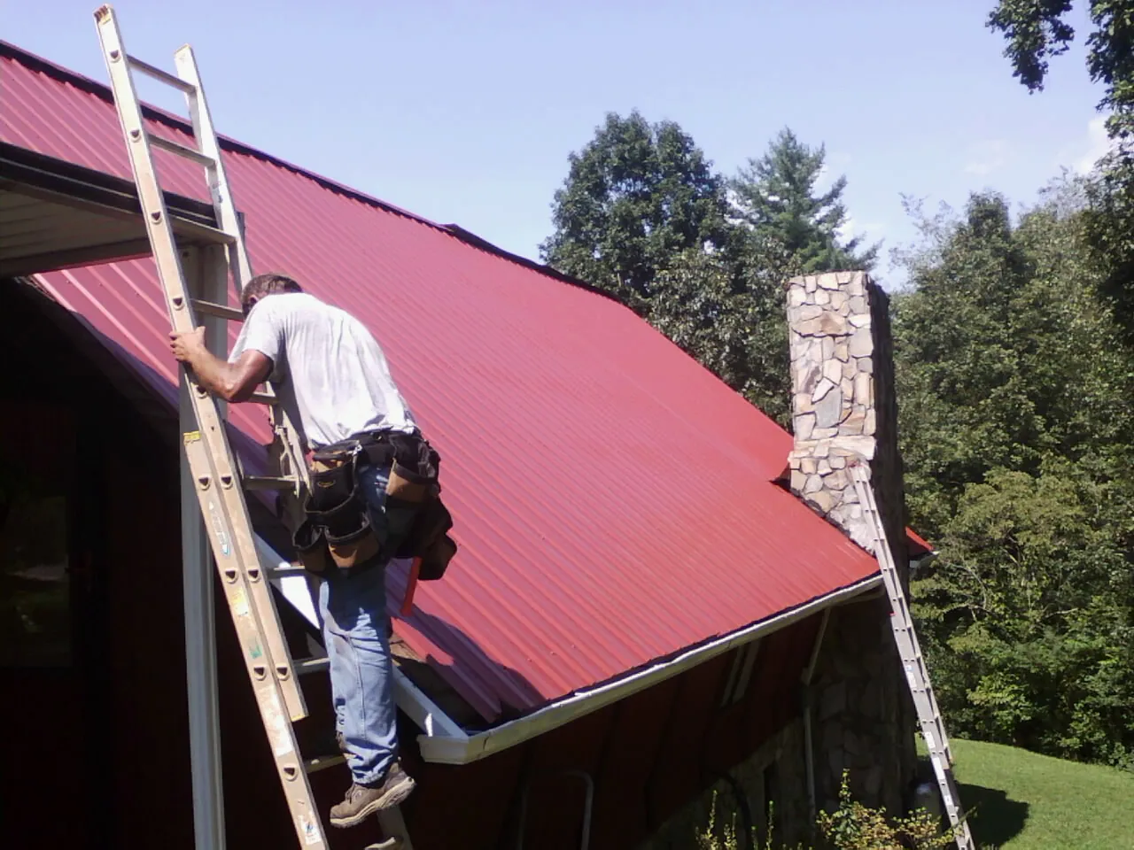 Roofer on a ladder beside a red metal roof and stone chimney — Soffit & Fascia Repair by Roofing Brothers in Hayfield